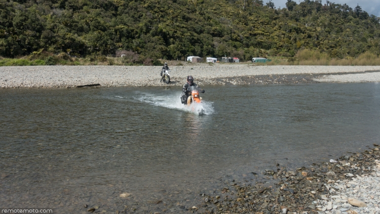 Motorbike and rider in the middle of fording a shallow river towards the camera, another bike and rider waiting on the far bank.